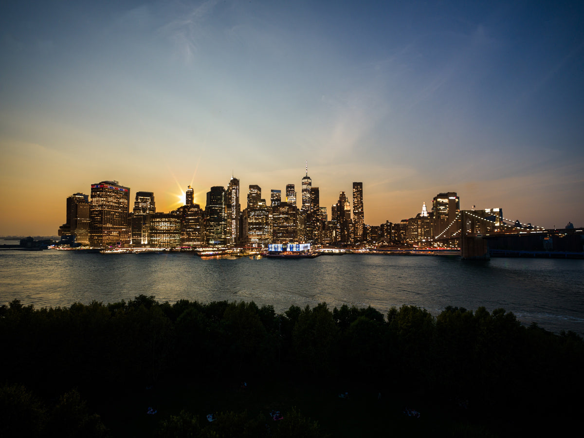 Warm early light on Manhattan’s skyline above a river and dark foreground foliage. Photograph by Anders Jorulf.