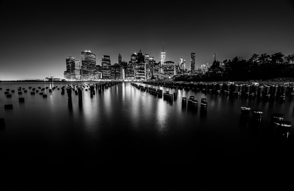 Black-and-white Manhattan skyline reflected in water behind old wooden pilings. Photograph by Anders Jorulf.