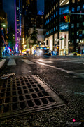 Low street-level New York night scene with coloured signs and a foreground drain cover. Photograph by Anders Jorulf.