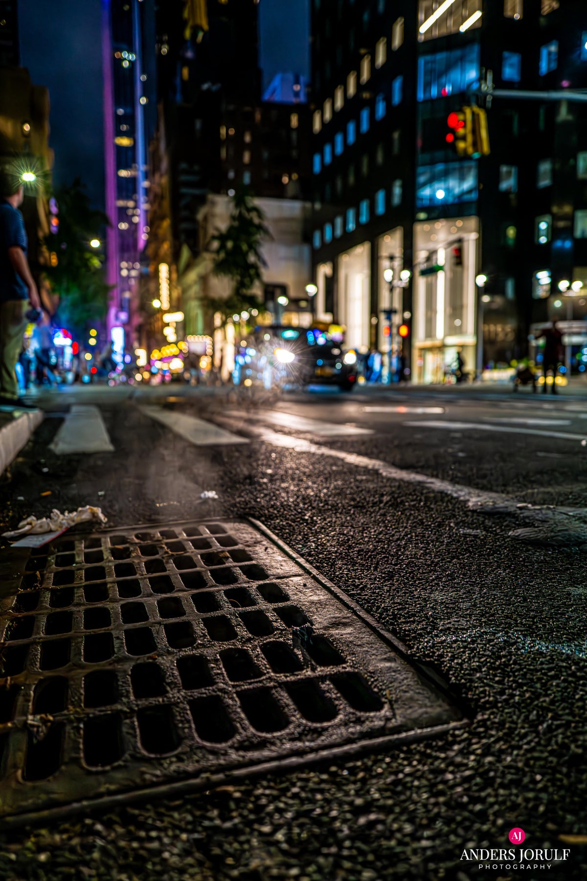 Low street-level New York night scene with coloured signs and a foreground drain cover. Photograph by Anders Jorulf.
