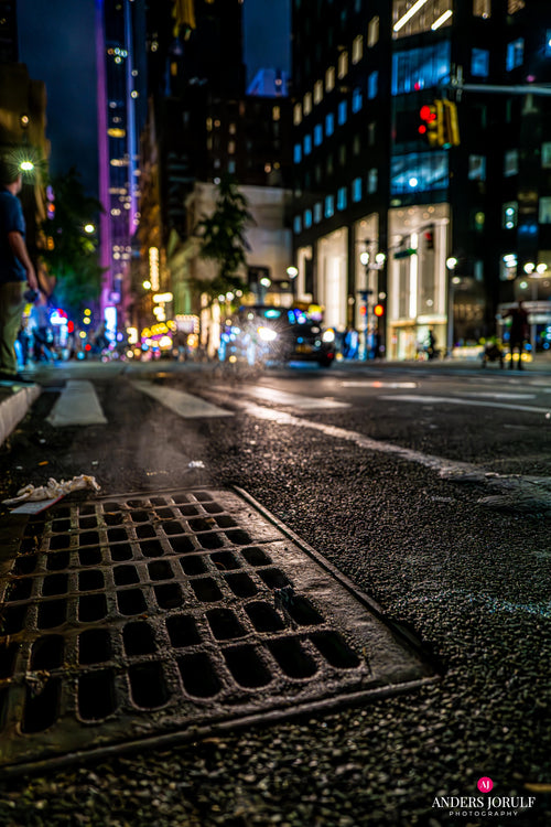 Low street-level New York night scene with coloured signs and a foreground drain cover. Photograph by Anders Jorulf.