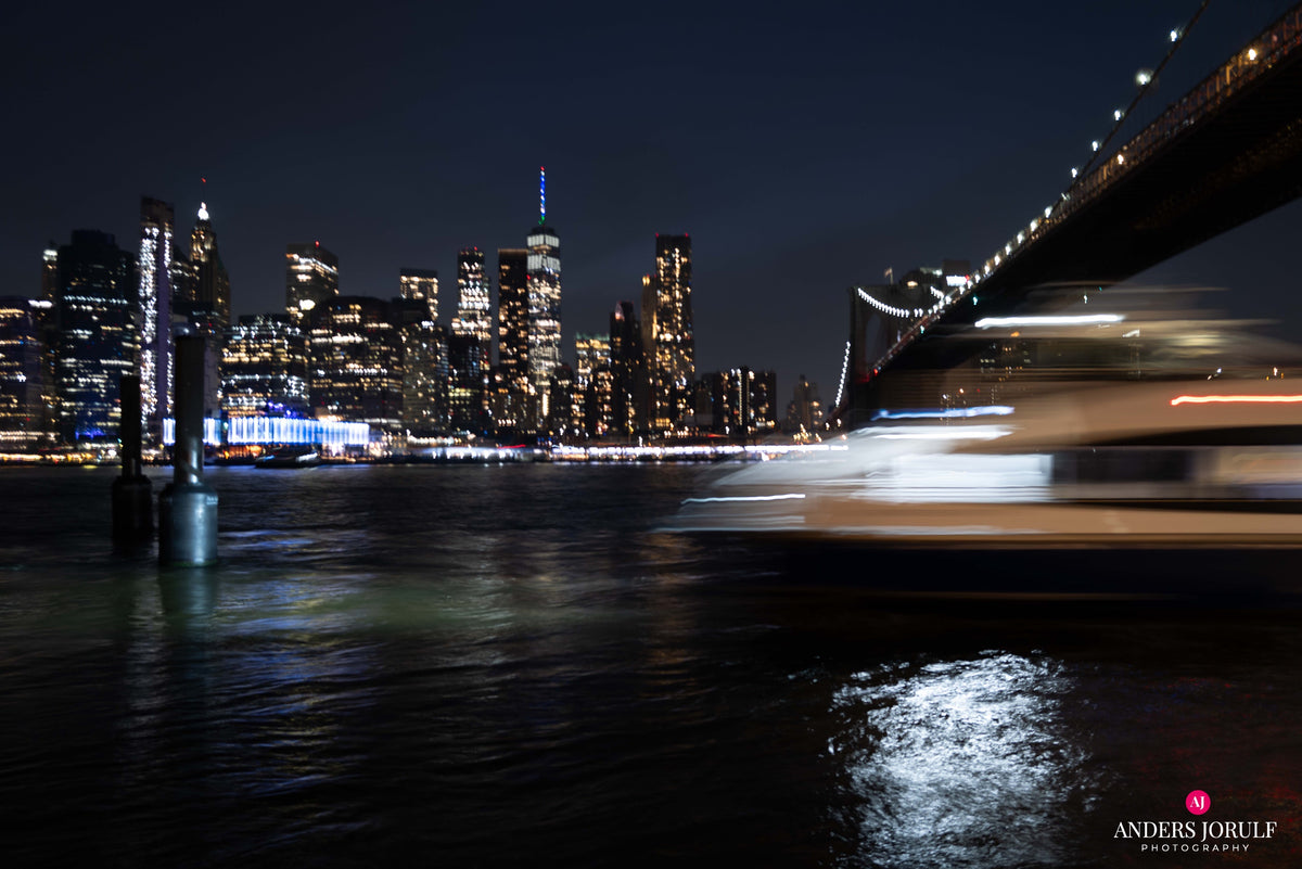 A boat passing the illuminated Manhattan skyline beside a bridge at night. Photograph by Anders Jorulf.